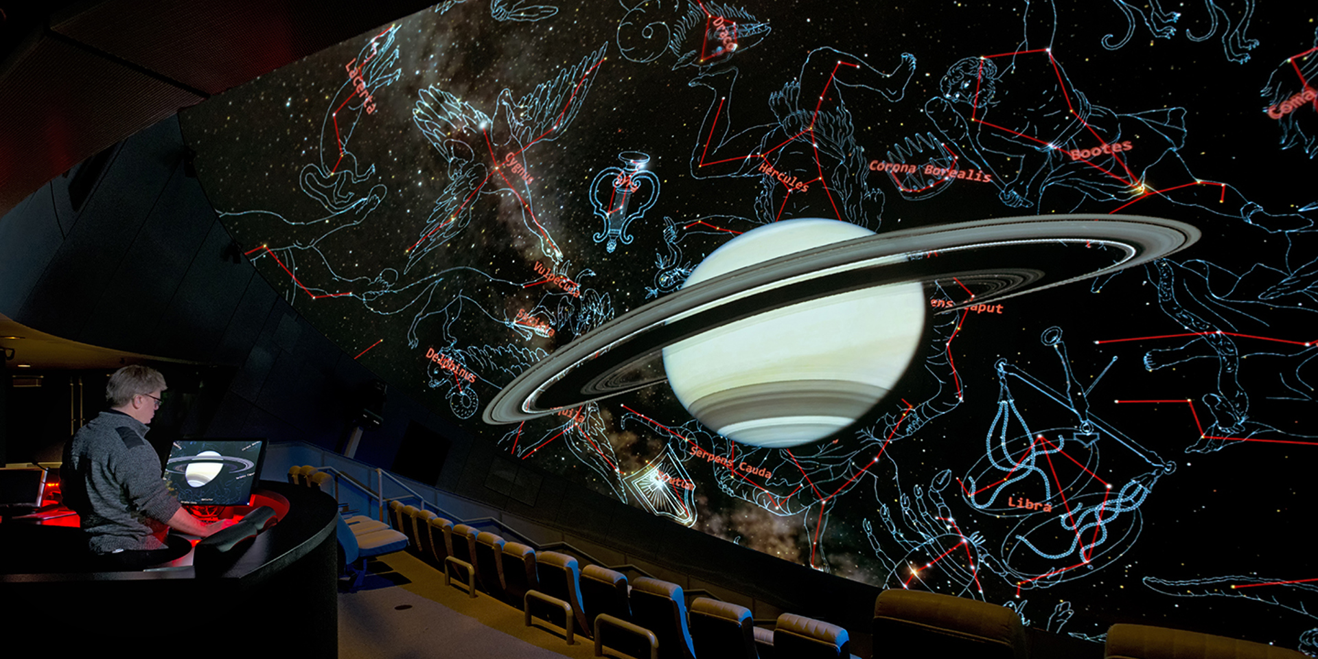 Image of a planetarium screen showing a ringed planet and constellations drawn around it, with a person sitting in the audience controlling the screen.