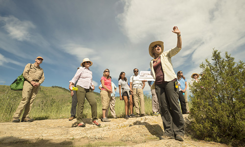 A group stands on a trail in a field listening to Michele Koons talk about the history of the Magic Mountain site.